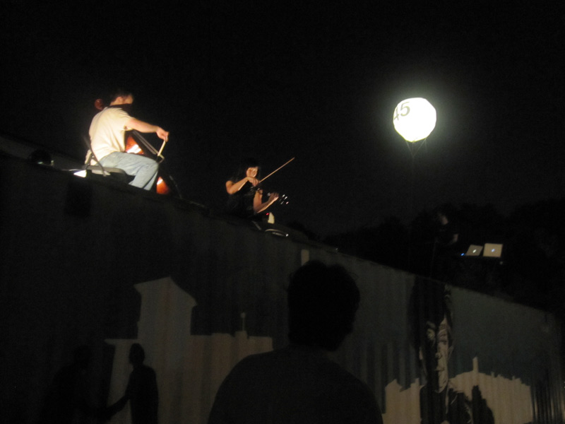 Matt Jarrard playing cello, Karyn Lu playing violin, and Matt Gilbert running two laptops on top of a storage container with a mural by Matt Hafner